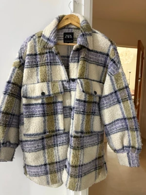 Rutig overshirt från ZARA i lila och beige - Säljer en fluffig overshirt från ZARA med rutigt mönster i lila, beige och vitt. Jackan har två stora bröstfickor med knappar, klassisk krage och avslappnad passform. Perfekt lager på lager och riktigt trendig just nu.