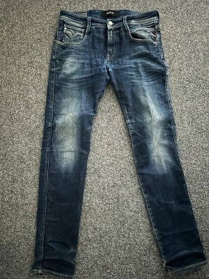 Blå slim fit jeans från Replay - Snygga blå slim fit jeans från Replay med slitningar och tvättade detaljer på benen. Klassisk femficksmodell med vita kontrastsömmar och Replay-logga på bakfickan. Jeansen har normal midja och är tillverkade i stretchigt denimtyg för extra komfort. Byxorna har ett hål i skrevet men det är enkelt at fixa med en symaskin eller hos en skräddare!