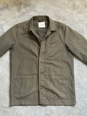 Olivgrön overshirt från A Day's March - Stilren olivgrön overshirt från A Day's March i bomull. Klassisk krage, tre utanpåliggande fickor och knäppning framtill. Perfekt lager-på-lager-plagg med avslappnad passform och snygga detaljer.
