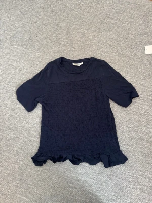 Mörkblå topp med volang från Gina Tricot - Supersöt mörkblå topp från Gina Tricot med smockad framsida och volangkant nedtill. Toppen har korta ärmar och en mjuk, bekväm känsla. Perfekt för dig som gillar en enkel men ändå detaljrik stil.