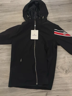 Svart vindjacka med huva Moncler -  Helt ny snygg svart vindjacka från Moncler med huva och hel dragkedja framtill. Jackan har en röd, vit och blå rand med Moncler-logga på ena ärmen. Tillverkad i lätt och tåligt material, perfekt för blåsiga dagar.