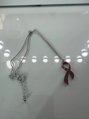 Halsband med rosa strassband - Snyggt halsband med ett hängsmycke i form av ett rosa band täckt av glittrande strass. Kedjan är silverfärgad och har justerbar längd. Perfekt för dig som vill ha ett statement-smycke med lite extra bling.