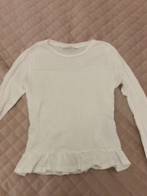 Vit långärmad topp med volang - Säljer en söt vit långärmad topp med smockad front och volangkant nedtill. Toppen har rund halsringning och är i ett mjukt material som känns bekvämt mot huden. Perfekt att styla med jeans eller kjol för en trendig look.