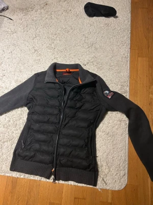 Mörkgrå quiltad cardigan med patch - Snygg mörkgrå cardiganjacka med quiltad front och stickade ärmar. Inga hål eller något, knappt användt sedan jag köpte den, limited edition parajumper 