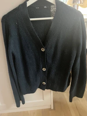 Svart kofta från 157 med knappar - Mysig svart kofta från 157 med tre stora knappar framtill. Koftan har v-ringning, lång ärm och är gjord i ett mjukt, lite luddigt material. Perfekt att slänga på sig över en t-shirt eller topp för en chill look.