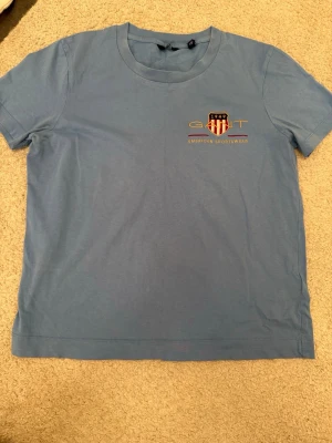 Blå kortärmad t-shirt från Gant - Säljer en ljusblå t-shirt från Gant med tryckt logga och sköld på bröstet. Klassisk rund halsringning och korta ärmar. Tillverkad i mjuk bomull som känns skön mot huden. Perfekt för en avslappnad och stilren look.