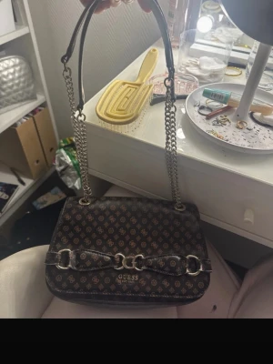 Brun monogramväska från Guess - Snygg brun handväska från Guess med klassiskt monogrammönster i beige. Väskan har silverfärgade kedjehandtag och dekorativa metalldetaljer framtill. Tillverkad i konstläder och har en stilren, rektangulär form. Perfekt accessoar för att lyfta din outfit.