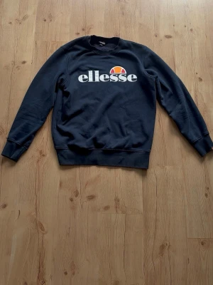 Mörkblå sweatshirt från Ellesse - Snygg mörkblå sweatshirt från Ellesse med stort vitt logotryck och färgglad detalj på bröstet. Tröjan har rund halsringning, ribbade muddar vid ärmslut och nederkant. Perfekt för en avslappnad och sportig stil.