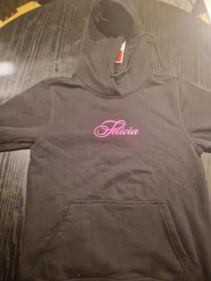 Svart hoodie med rosa tryck - Säljer en svart hoodie med huva och magficka. På bröstet står det 'Felicia' i rosa skrivstil och på ryggen finns ett stort rosa tryck av en ros. Hoodien har klassisk passform och är gjord i mjukt bomullsmaterial.