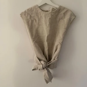 Beige topp med knytning framtill - Stilren beige topp i ett strukturerat tyg med rund halsringning och korta ärmar. Toppen har en snygg knytning framtill som ger en avslappnad men trendig look. Perfekt att matcha med jeans eller kjol för en enkel men cool outfit.