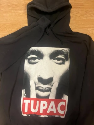 Svart Tupac hoodie från Pull&Bear - Svart hoodie med stor svartvit Tupac-print på framsidan och röd/vit text. Klassisk huva med dragsko och mjukt material. Perfekt för dig som gillar streetwear och vill ha en unik look.
