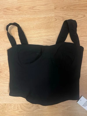 Svart korsett-topp från Boohoo - Trendig svart korsett-topp från Boohoo med breda axelband och markerade kupor. Toppen är croppad och har en stilren, figurnära passform som passar perfekt till höga jeans eller kjol.