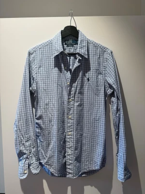 Blårutig skjorta från Polo Ralph Lauren - Klassisk blårutig skjorta från Polo Ralph Lauren i slim fit-modell. Skjortan har långa ärmar, vit knappstängning och en liten broderad logga på bröstet. Tillverkad i mjukt bomullstyg och passar perfekt till jeans eller chinos.