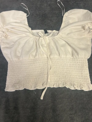 Vit croppad linnetopp med knyt - Söt vit croppad linnetopp med tunna axelband och smockad nederdel. Toppen har knyt framtill och lätt puffade detaljer vid axlarna för en trendig look. Perfekt till sommaren och enkel att matcha med jeans eller kjol.