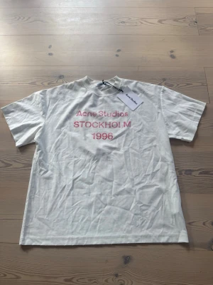 Vit Acne Studios t-shirt Stockholm - Vit t-shirt från Acne Studios med tryck i rosa på bröstet där det står 'Acne Studios STOCKHOLM 1996'. Klassisk rak passform och rund hals. Tillverkad i mjuk bomull och har korta ärmar. Perfekt för en clean och stilren look. Aldrig använd 