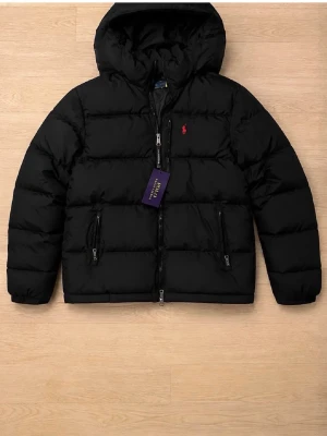 Svart pufferjacka med huva - Snygg svart pufferjacka från Polo Ralph Lauren med huva och två dragkedjeförsedda fickor. Jackan har en klassisk quiltad design och den ikoniska röda loggan på bröstet. Perfekt för kalla vinterdagar och riktigt skön att bära.