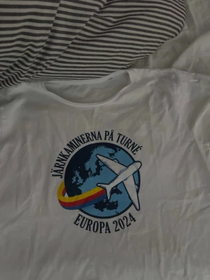 Vit t-shirt Järnkaminerna Europa 2024 - Vit t-shirt med tryck av ett flygplan och texten 'Järnkaminerna på turné Europa 2024'. Färgglad regnbågsdetalj och motiv av en jordglob framtill. Klassisk passform och rund halsringning. Perfekt för dig som gillar supporterkläder och vill sticka ut.