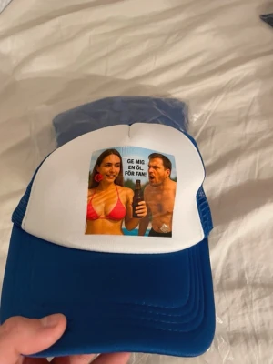 Blå och vit truckerkeps med meme-tryck - Säljer en blå och vit truckerkeps med mesh på sidorna och justerbar snapback. Framtill finns ett färgglatt meme-tryck med texten 'GE MIG EN ÖL, FÖR FAN!'. Perfekt för dig som gillar roliga accessoarer och vill sticka ut.