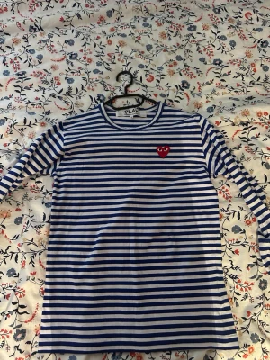 Randig långärmad tröja Comme des Garçons - Säljer en blåvit randig långärmad tröja från Comme des Garçons Play med det ikoniska röda hjärtat med ögon broderat på bröstet. Tröjan har rund halsringning och är gjord i mjuk bomull. Perfekt för en chill och stilren look.
