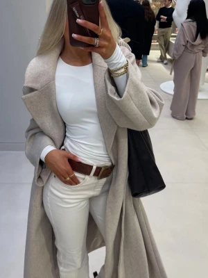 Beige kappa med knytskärp - Stilren beige kappa i mjukt material med bred krage och knytskärp i midjan. Lång modell som ger en avslappnad men ändå elegant look. Perfekt att slänga över en outfit för en trendig vibe.