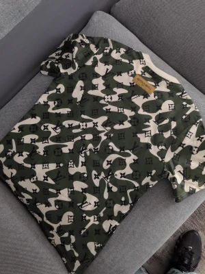 Camouflage t-shirt - Säljer en grön och vit t-shirt med ett unikt camouflage-mönster och loggor. T-shirten har korta ärmar och rund halsringning. Perfekt för dig som vill sticka ut med en exklusiv och trendig look. Passar även M.