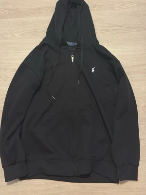 Svart hoodie från Polo Ralph Lauren - Svart hoodie med dragkedja från Polo Ralph Lauren. Klassisk design med huva och dragsnören, samt fickor framtill. Ikoniska vita broderade loggan på bröstet. Perfekt för en avslappnad och stilren look.