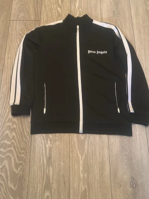 Svart track jacket från Palm Angels - Svart track jacket från Palm Angels med vita ränder längs ärmarna och vit dragkedja framtill. Tröjan har hög krage, två fickor med dragkedja och logga på bröstet. Perfekt för en sportig och trendig look.