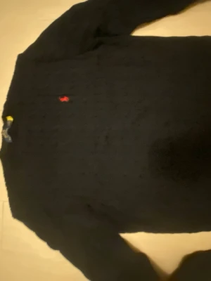 Mörkblå Ralph Lauren kabelstickad tröja - Snygg mörkblå kabelstickad tröja med rund hals och lång ärm. Tröjan har ett litet rött broderat märke på bröstet och är gjord i ett mjukt stickat material. Perfekt för dig som gillar klassisk och stilren look.