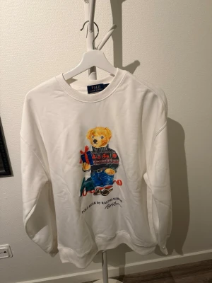 Vit sweatshirt Polo bear - Vit sweatshirt från Polo Ralph Lauren med en ikonisk björn på framsidan som har på sig en stickad tröja och jeans. Rund halsringning, ribbade muddar och avslut. Mjuk bomullskänsla och avslappnad passform. Perfekt för dig som gillar klassiska streetwear-vibbar. Säljer då den va för stor för mig. Den är alltså helt ny.
