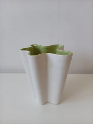 Vit och grön vågig vas i keramik - Säljer en unik vas i keramik med vågig form. Utsidan är vit och insidan har en ljusgrön färg. Perfekt som dekoration eller för blommor. Formen är organisk och modern, vilket gör den till ett coolt inslag i rummet. Den ä lite smudig där inne som ja int flrlskt få bort 