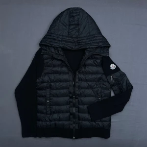 Svart Moncler cardigan med huva  - Snygg svart cardigan från Moncler med huva och stickade svarta ärmar. Cardiganen har dragkedja framtill, två sidofickor, Moncler-logga på vänster ärm och fungerande NFC märke. Materialet är en mix av dun och stickat tyg som ger en cool kontrast.