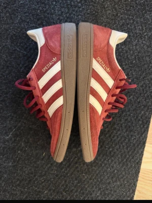 Adidas Spezial vinröda sneakers - Adidas Spezial sneakers i vinrött mocka med klassiska vita ränder på sidorna och bruna gummisulor. Skorna har röda skosnören och vit detalj vid hälen. Perfekta för dig som gillar retrostil och streetwear.