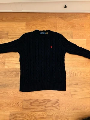 Polo Ralph Lauren cableknit  - Mörkblå kabelstickad tröja från Polo Ralph Lauren med rund halsringning och röd broderad logga på bröstet. Tröjan har långa ärmar och ribbade muddar vid ärmslut och nederkant. Perfekt för en stilren och klassisk look.