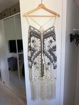 Vit långklänning med fransar och mönster - Säljer en vit klänning med svarta broderade mönster och tunna axelband. Klänningen har dekorativa fransar längst ner och ett bohemiskt utseende med snygga detaljer. Perfekt för sommarens alla tillfällen! Från märket Odd Molly i storlek 3 som enligt deras storlekguide motsvarar en storlek L. Helt ny med tags/ prislapp fortfarande kvar. 
