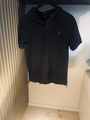 Svart Ralph Lauren piké  - Klassisk svart Ralph lauren piké  med röd broderad logga på bröstet. Tillverkad i mjuk bomull och har en krage med knappar framtill. Perfekt för en avslappnad men ändå stilren look. Storlek L men passar M, pris kan diskuteras vid snabb affär