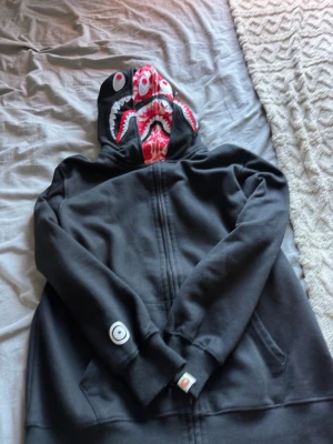 Svart hoodie med hajmotiv på huvan - Cool svart hoodie med dragkedja och unik huva som har ett hajmotiv med ögon och tänder i rött, vitt och svart. Hoodien har fickor framtill och ribbade muddar vid ärmar och nederkant. Perfekt för dig som vill sticka ut med din stil.