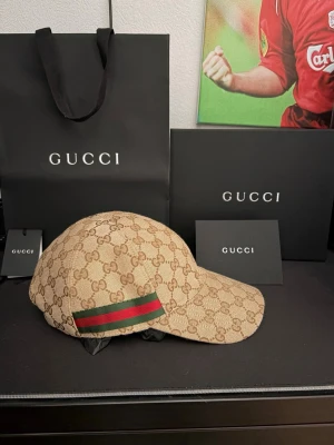 Beige keps från Gucci med GG-mönster - Kepsen är helt ny och köpt för två månader sedan. Säljer en beige keps från Gucci med det klassiska GG-monogrammet över hela kepsen. På sidan finns en grön och röd rand som signaturdetalj. Kepsen har böjd skärm och justerbar rem bak. Perfekt för dig som vill ha en exklusiv och trendig accessoar. 