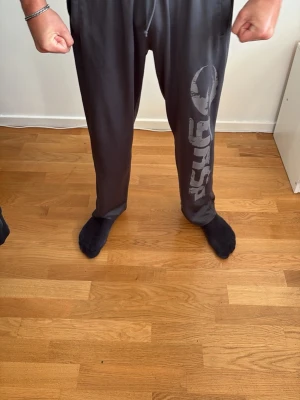Gasp byxor - Säljer ett par mörkgrå gymbyxor med stort tryck längs ena benet. Byxorna har dragsko i midjan och är tillverkade i ett mjukt, stretchigt material som passar perfekt för chill dagar. Snygg och avslappnad passform.