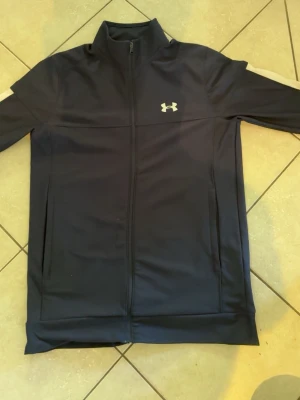 Marinblå zip från Under Armour - Snygg marinblå zip från Under Armour med hel dragkedja framtill och hög krage. Jackan har två sidofickor och ett vitt logotryck på bröstet. Tillverkad i ett lätt och ventilerande material som passar perfekt till träning.