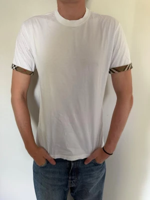 Vit Burberry T-shirt  - Vit t-shirt från Burberry med klassiska rutiga detaljer på ärmsluten. T-shirten har rund halsringning och korta ärmar. Köpt i Milano för 2000k med kvitto i bilderna. Nypris ca 3000kr. Storlek M men skulle säga att den är lite liten i storleken, närmare S. Använd enstaka gånger. Hör av dig om du har några frågor eller vill ha fler bilder. 