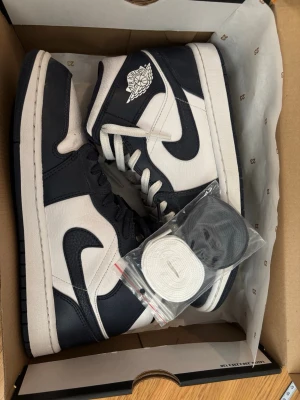 Air Jordan 1 Mid Obsidian/Vit - Snygga Air Jordan 1 Mid sneakers i färgerna obsidianblå och vit. Klassisk design med ikonisk vinge-logga på sidan och svart swoosh. Skorna är i läder och kommer med extra skosnören i både vitt och svart.