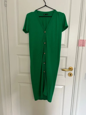 Grön ribbad klänning från Zara - Snygg grön ribbad klänning från Zara med v-ringning och knappar framtill. Klänningen har korta ärmar och är i en mjuk, stretchig polyesterblandning. Perfekt för dig som vill ha en enkel men trendig look.