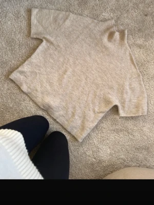 Beige stickad tröja från Gina Tricot - Kortärmad stickad tröja i beige från Gina Tricot. Tröjan har hög krage och är gjord i en mjuk ullblandning. Perfekt för lager på lager och enkel att matcha med olika outfits. Har en fläck på helsen men tror det går bort i tvätten 