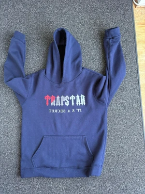 Mörkblå Trapstar hoodie med tryck - Snygg mörkblå hoodie med stor huva och magficka. På bröstet står det 'TRAPSTAR' i vitt och rött samt texten 'IT'S A SECRET' i vitt. Hoodien har en klassisk passform och är gjord i mjukt sweatshirtmaterial. Perfekt för en streetwear-look.