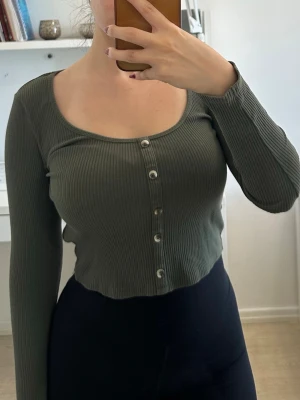 Olivgrön ribbad croppad topp - Säljer en olivgrön ribbad topp med lång ärm och croppad passform. Toppen har rundad urringning och knappar framtill som detalj. Perfekt att styla med höga jeans eller kjol för en trendig look.