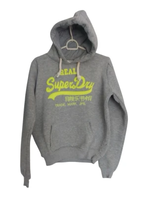 Grå Superdry hoodie med gul text - Snygg grå hoodie från Superdry med stor gul logga och text på bröstet. Klassisk känguruficka framtill och vita dragsnören i huvan. Perfekt för en avslappnad och sportig stil. Mjuk bomullsblandning som känns skön mot huden.