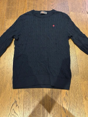 Mörkblå kabelstickad tröja - Kabelstickad mörkblå tröja med rund hals och lång ärm. Tröjan har ett diskret rött broderat emblem på bröstet och ribbade muddar vid ärmslut och nederkant. Perfekt för lager på lager och en klassisk look. Ett litet hål nere på vänster sida av mage!