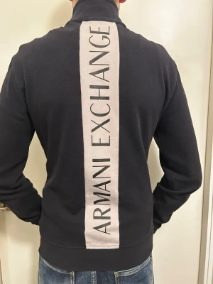 Svart tröja från Armani Exchange med dragkedja - Säljer en svart jacka från Armani Exchange med hel dragkedja framtill och stor logga längs ryggen. Jackan har lång ärm och är tillverkad i en mjuk bomullsblandning med inslag av polyester och elastan för extra komfort.