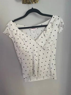 Vit prickig topp från Gina Tricot - Söt vit topp från Gina Tricot med svarta prickar och v-ringad hals. Toppen har korta ärmar med små volangdetaljer och är gjord i mjuk bomull. Perfekt för dig som gillar en feminin och lekfull stil.