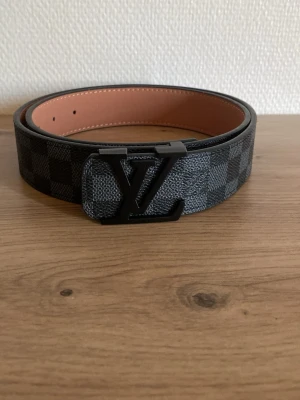 Svart och grått rutigt  bälte - Snyggt bälte med svart och grått rutmönster och stor svart metall -logga i metall framtill. Bältet har en klassisk design och insidan är beige. Perfekt accessoar för att lyfta din outfit.